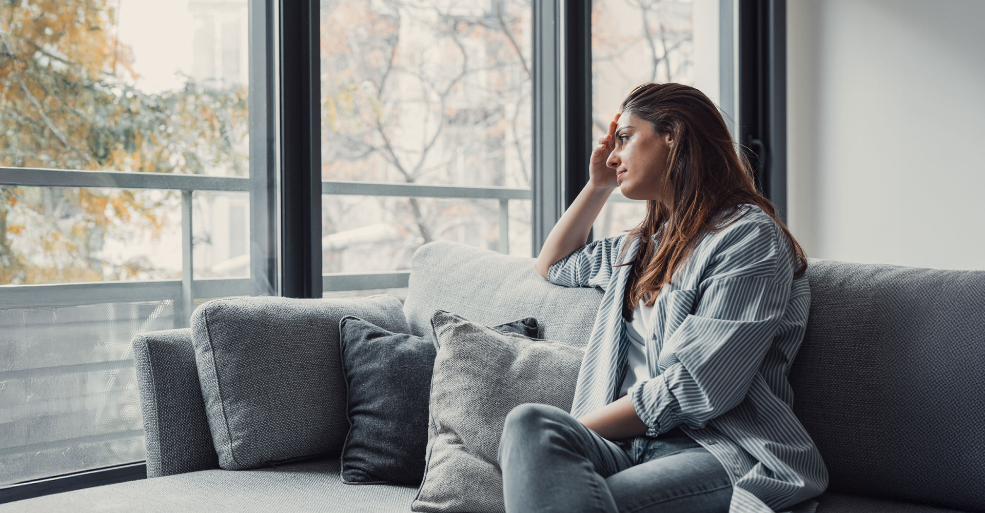 Frau sitzt nachdenklich auf einem Sofa und schaut aus dem Fenster, den Kopf mit der Hand stützend.