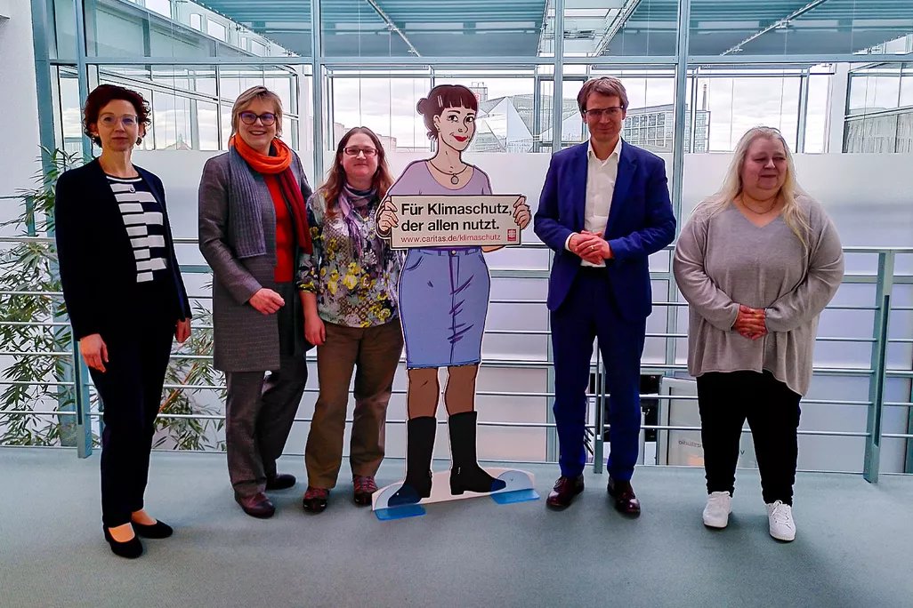 Gruppe steht neben Klimaschutz-Figur ‚Jenny‘ mit Schild ‚Für Klimaschutz, der allen nutzt‘ in einem Innenbereich