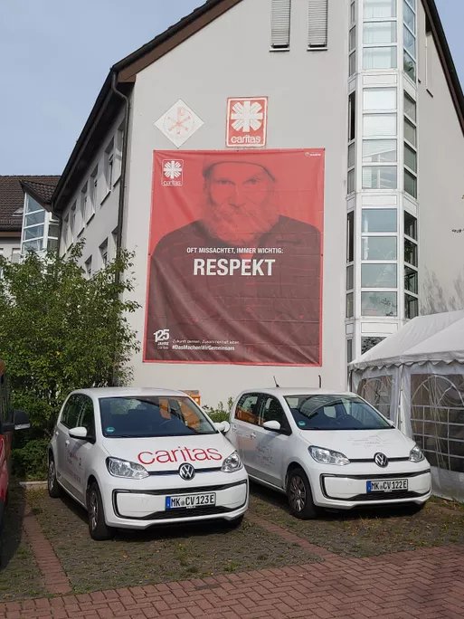Caritas-Gebäude mit großem Banner ‚Respekt‘ und zwei Caritas-Fahrzeugen vor dem Eingang