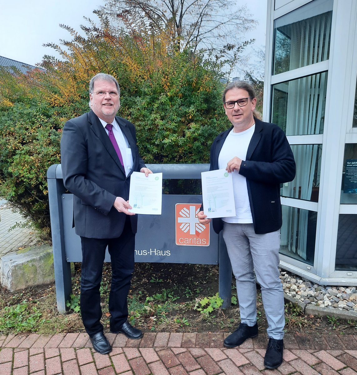 Zwei Personen überreichen Zertifikate vor einem Caritas-Schild am Eingang einer Einrichtung