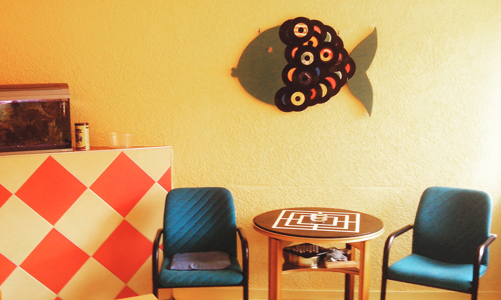 Gemütlicher Aufenthaltsraum mit zwei blauen Stühlen, einem kleinen Holztisch mit Spielbrett, einem Aquarium und einer Fisch-Wanddekoration aus bunten runden Elementen.